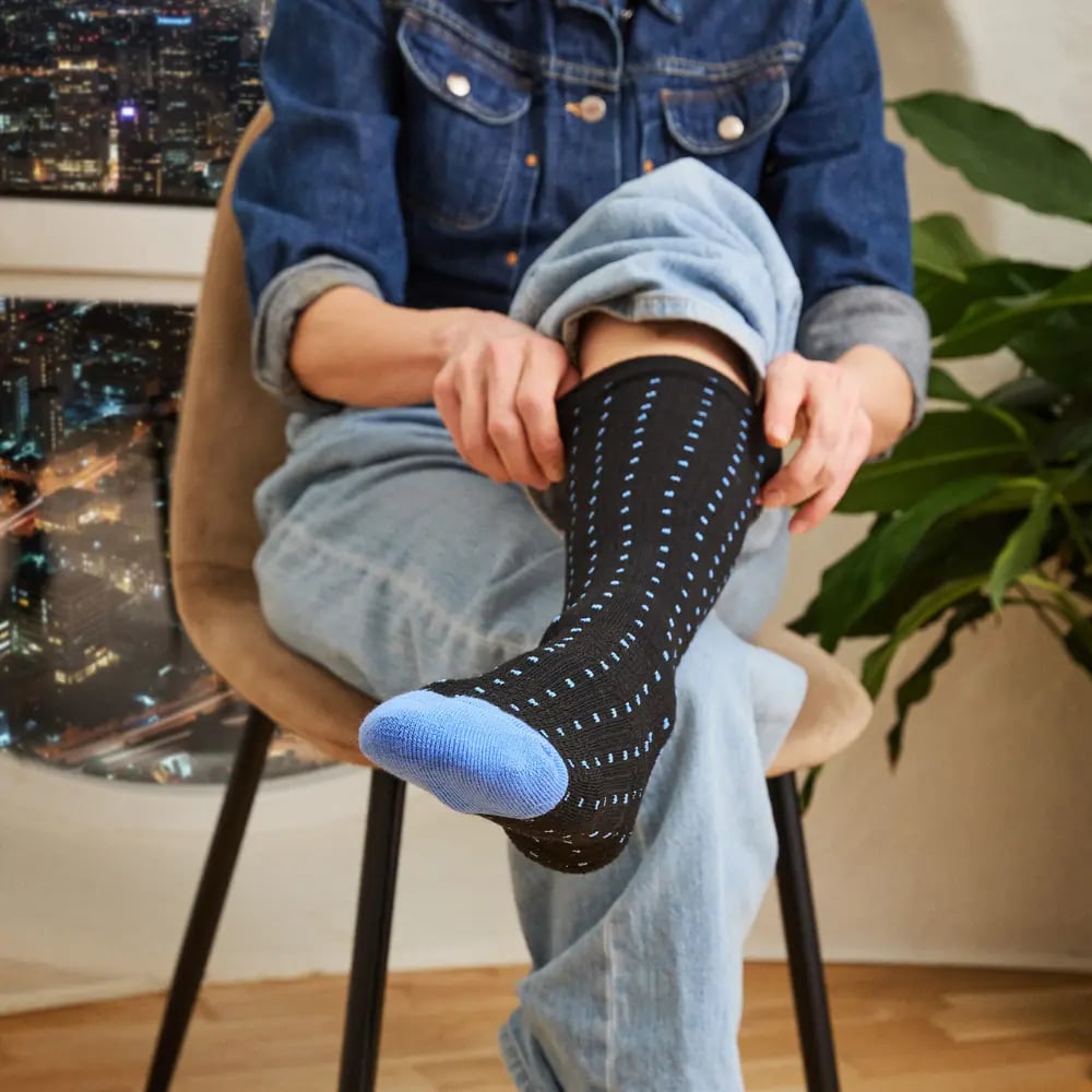A person wearing jeans sits in a chair and pulls on a black sock with a blue pattern.